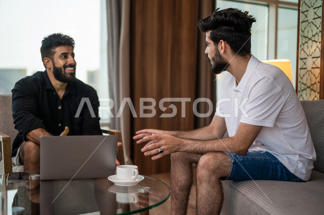 شابان عربيان خليجيان سعوديان مبتسمان يجلسان في المقهى، الاستمتاع بشرب القهوة الساخنة، استخدام جهاز الحاسوب المحمول، حوار ونقاش، كوب قهوة، مشروبات ساخنة، ديوانية شباب السعودية، جلسة صداقة، أجواء شبابية في مقهى سعودي