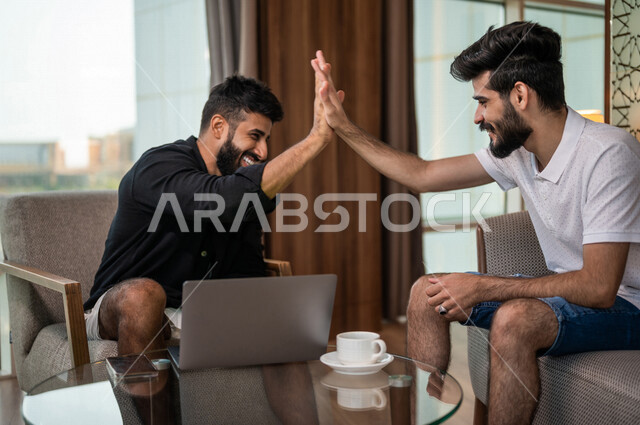 شابان عربيان خليجيان سعوديان مبتسمان يجلسان في المقهى، الاستمتاع بشرب القهوة الساخنة، استخدام جهاز الحاسوب المحمول، حوار ونقاش، كوب قهوة، مشروبات ساخنة، ديوانية شباب السعودية، جلسة صداقة، أجواء شبابية في مقهى سعودي