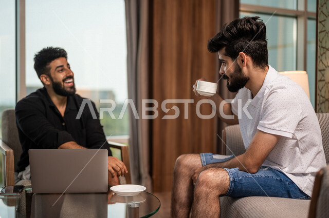 شابان عربيان خليجيان سعوديان مبتسمان يجلسان في المقهى، الاستمتاع بشرب القهوة الساخنة، استخدام جهاز الحاسوب المحمول، حوار ونقاش، كوب قهوة، مشروبات ساخنة، ديوانية شباب السعودية، جلسة صداقة، أجواء شبابية في مقهى سعودي
