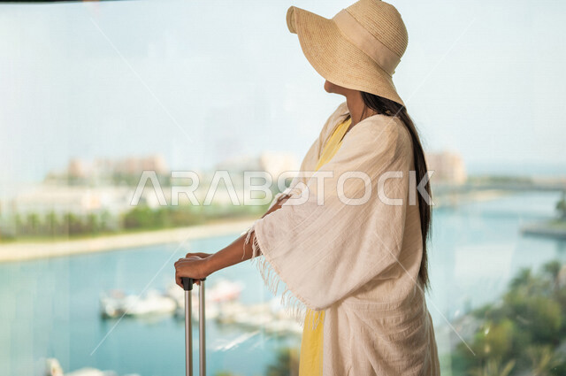 Close-up of a Saudi Arabian Gulf woman on a tourist trip, a window overlooking the waterfront in Jeddah, wearing a summer casual outfit, a travel bag, preparing for the summer vacation, a hotel resort, fun summer activities