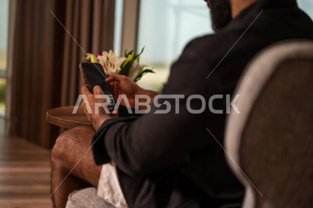 صورة مقربة لشاب عربي خليجي سعودي يرتدي زي كاجوال صيفي ويقوم باستخدام الهاتف المحمول، طلبات وتسوق اونلاين، حجوزات فندقية، تحديد الوجهات السياحة، نشاطات ترفيهية، الدردشة والتواصل الاجتماعي، تصفح المواقع الالكترونية، التواصل الاجتماعي
