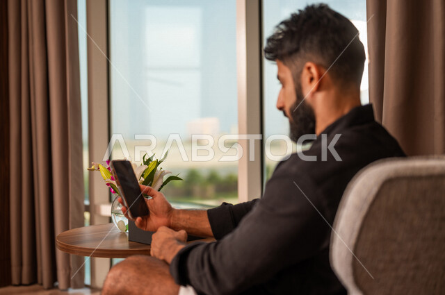 صورة مقربة لشاب عربي خليجي سعودي يرتدي زي كاجوال صيفي ويقوم باستخدام الهاتف المحمول، طلبات وتسوق اونلاين، حجوزات فندقية، تحديد الوجهات السياحة، نشاطات ترفيهية، الدردشة والتواصل الاجتماعي، تصفح المواقع الالكترونية، التواصل الاجتماعي