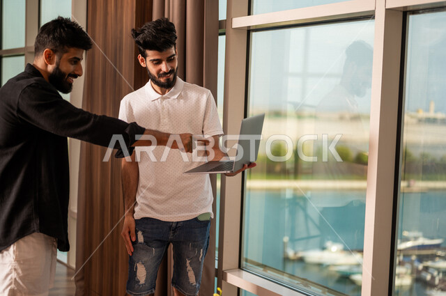 شابان عربيان خليجيان سعوديان يستخدمان جهاز الحاسوب المحمول، التسوق عبر الانترنت، بث مباشر، أنشطة شبابية، التخطيط للعطلة الصيفية، حجوزات فندقية، تحديد وجهات سياحية، زي كاجوال، اطلالة بحرية، قضاء وقت ممتع، مصيف فندقي سياحي