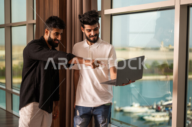 شابان عربيان خليجيان سعوديان يستخدمان جهاز الحاسوب المحمول، التسوق عبر الانترنت، بث مباشر، أنشطة شبابية، التخطيط للعطلة الصيفية، حجوزات فندقية، تحديد وجهات سياحية، زي كاجوال، اطلالة بحرية، قضاء وقت ممتع، مصيف فندقي سياحي