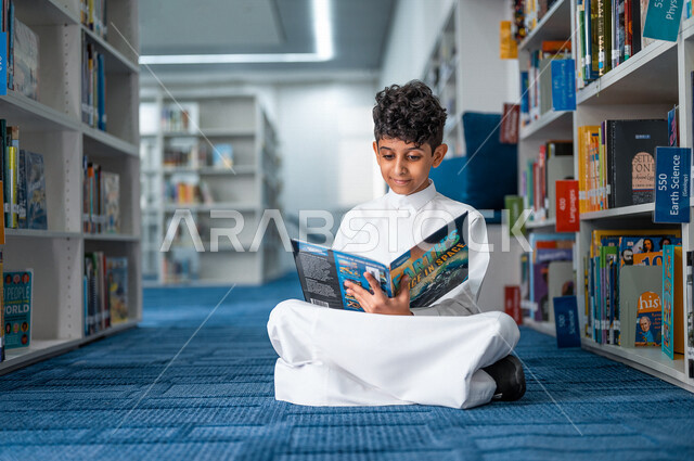 طالب عربي سعودي خليجي يقوم بقراءة كتاب من المكتبة المدرسية، الاستمتاع بقراءة الكتب، مفهوم القراءه والثقافه, مكتبة المدرسة، التعليم الذاتي, بيئه مدرسيه تعليميه متطوره, مدارس أكاديميه في المملكة العربية السعودية