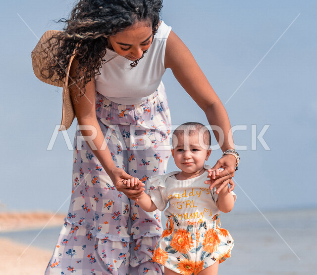 صورة مقربة لأم عربية خليجية سعودية تمسك إبنتها بحب وحنان، المشي على رمل شاطئ البحر، الاستمتاع بالاجازة الصيفية، مفهوم الأمومة والطفولة، قضاء أوقات عائلية ممتعة بالعطلة الصيفية على شاطئ بحر جدة السياحي