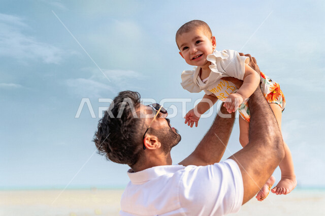 صورة مقربة لأب عربي خليجي سعودي يرتدي النظاراة الشمسية ويحمل إبنته بحب وحنان، إيماءات تدل على السعادة و المحبة، أجواء عائلية سعيدة، مفهوم الأبوة والطفولة، قضاء أوقات عائلية ممتعة بالعطلة الصيفية على شاطئ بحر جدة السياحي