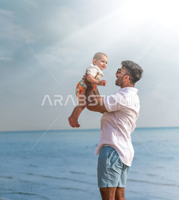 صورة مقربة لأب عربي خليجي سعودي يرتدي النظاراة الشمسية ويحمل إبنته بحب وحنان، إيماءات تدل على السعادة و المحبة، أجواء عائلية سعيدة، مفهوم الأبوة والطفولة، قضاء أوقات عائلية ممتعة بالعطلة الصيفية على شاطئ بحر جدة السياحي