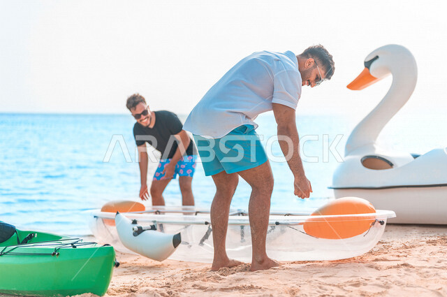 صديقان عربيان خليجيان سعوديان يرتدون النظارة الشمسية ويقضيان وقت ممتع سويا على بحر جدة السياحي، قارب التجديف البلاستيكي الشفاف، الاستعداد لركوب زورق التجديف، أنشطة ترفيهية شبابية، المتعة بين الأصدقاء، هوايات ورياضات شبابية، سياحة وإستجمام، العطلة الصيفية