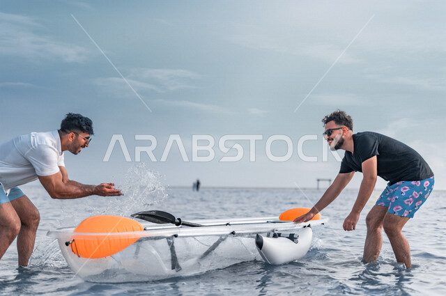 صديقان عربيان خليجيان سعوديان يرتدون النظارة الشمسية ويقضيان وقت ممتع سويا على بحر جدة السياحي، قارب التجديف البلاستيكي الشفاف، الاستعداد لركوب زورق التجديف، أنشطة ترفيهية شبابية، المتعة بين الأصدقاء، هوايات ورياضات شبابية، سياحة وإستجمام، العطلة الصيفية
