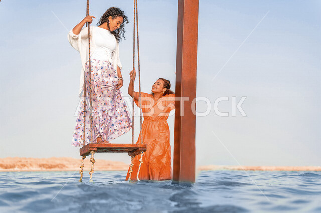 صديقتان عربيتان خليجيتان سعوديتان تقضيان وقت ممتع على الارجوحة المعلقة فوق البحر، بحر جدة السياحي، انشطة ترفيهية، الاستمتاع بالاجازة الصيفية، ايماءات وجه ويدين تدل على السعادة، سياحة وإستجمام، انشطة ترفيهية، العطلة الصيفية، أزياء صيفية