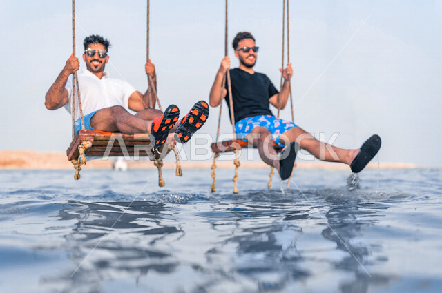 صديقان عربيان خليجيان سعوديان يقضيان وقت ممتع على الارجوحة المعلقة فوق البحر، بحر جدة السياحي، انشطة ترفيهية، الاستمتاع بالاجازة الصيفية، ايماءات وجه ويدين تدل على السعادة، سياحة وإستجمام، انشطة ترفيهية، العطلة الصيفية، أزياء صيفية