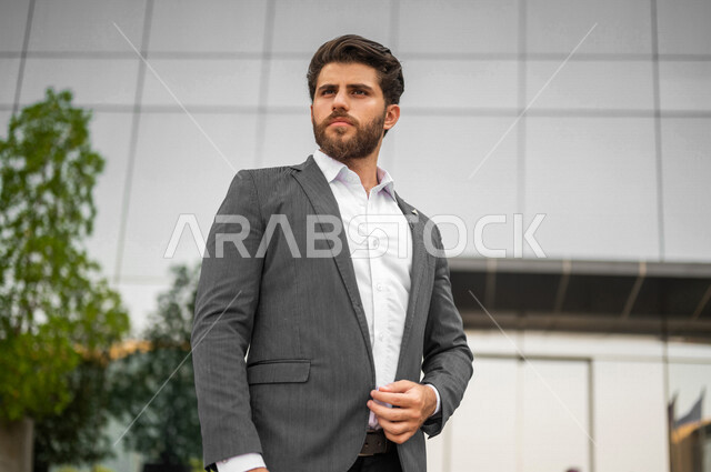 صوره مقربه لرجل أعمال عربي يرتدي البدله الرسميه يقف خارج مبنى الشركة، الإنتظار في الخارج,إيماءات تدل على التفكير, النظر و التأمل, إنتهاء الدوام، خروج الموظفين، مهن ووظائف سعودية، شركات ومكاتب خليجيه سعوديه في المملكه العربيه السعوديه