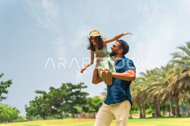 رجل عربي خليجي سعودي يقضي وقت ممتع مع إبنته في إحدى الحدائق الترفيهية بالسعودية، فعاليات ترفيهية عائلية، عطلة صيفية، حدائق ونزهات ترفيهية للعائلة في المملكة العربية السعودية، أجواء عائلية سعيدة، الأب يحمل إبنته إلى الأعلى، تعابير تدل على السعادة