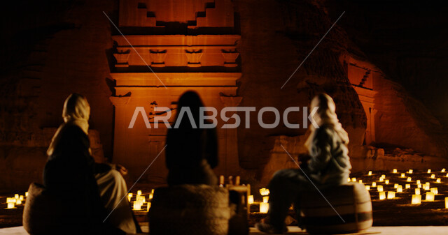 A picture from the back of Saudi Arabian Gulf women sitting in front of the Flying Museum in Al-Ula, Saudi Arabia, lit candles, Winter at Tantora festival in the historic stone in Al-Ula, tourism in Saudi Arabia