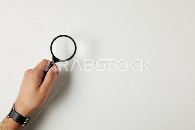 صورة مقربة ليد رجل يحمل عدسة مكبرة، استخدام العدسة المكبرة، الفحص والإستكشاف، رؤية عن قرب، التدقيق والتركيز، تكبير الاشياء، أدوات بصرية، خلفية بيضاء