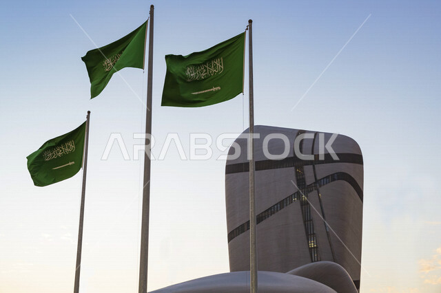 محموعة من اعلام المملكة العربية السعودية ترفرف ومن خلفها مركز الملك عبدالعزيز الثقافي العالمي (إثراء) في مدينة الظهران في المملكة العربية السعودية، سارية العلم، معالم ثقافية مشهورة، الفن الهندسي المعماري، جمال مبنى مركز الملك عبد العزيز، السياحة في السعود