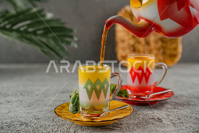 صورة مقربة لابريق الشاي مع الاكواب الملونة، سكب مشروب الشاي الساخن في الكوب، مشروبات دافئة، ابريق شاي، اكواب زجاجية مزخرفة، اوراق النعناع الخضراء