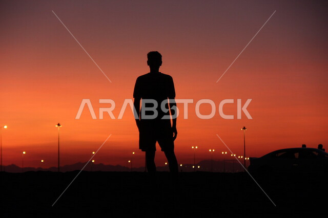 صورة ظلية لشاب يقف في الشارع اثناء غروب الشمس، غروب الشمس الساحر، اعمدة الانارة، جمال السماء الحمراء وقت الغروب