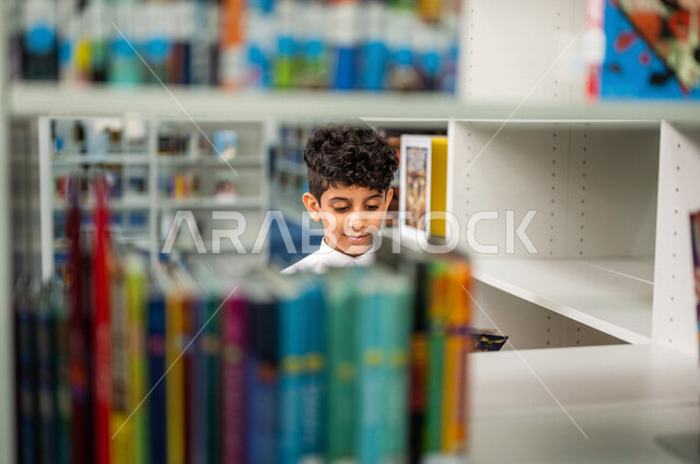 طالب عربي سعودي خليجي يقوم بإختيار كتاب من المكتبة المدرسية، الاستمتاع بقراءة الكتب، مفهوم القراءه والثقافه, مكتبة المدرسة، التعليم الذاتي, بيئه مدرسيه تعليميه متطوره, مدارس أكاديميه في المملكة العربية السعودية
