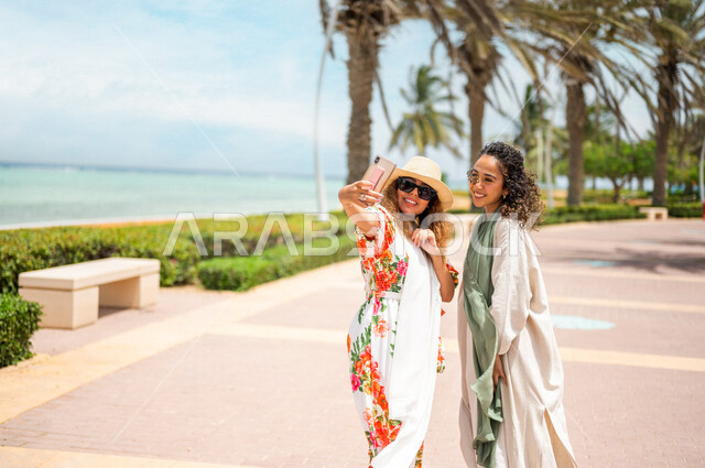صديقتان عربيتان سعوديتان خليجيتان تلتقطان صور تذكاريه في ممشى كورنيش جده, إستخدام الهاتف المحمول, شاطيء سياحي, التنزه في جده, دردشة ومحادثات, الإستمتاع مع الأصدقاء, الواجهه البحريه لمدينة جده, عطله صيفيه