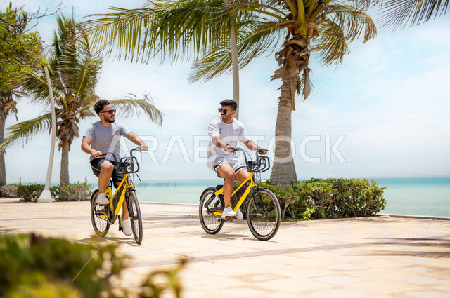 Two Saudi Arabian Gulf youths riding a bicycle in the Jeddah Corniche, the waterfront of the city of Jeddah, a tourist beach, cycling, practicing entertainment on the Jeddah Corniche promenade, sharing recreational activities with friends, tourism in Saud