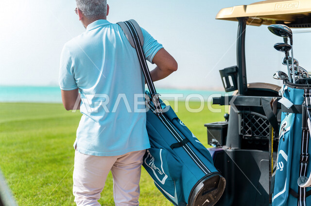 زاوية من الخلف لرجل عربي خليجي سعودي يرتدي الزي الرياضي ويحمل حقيبة الجولف، ممارسة لعبة الغولف على مساحات واسعة خضراء، أنشطة رياضية بالسعودية، لعبة الجولف، رياضات الألعاب الأولمبية الصيفية، أنشطة ترفيهية
