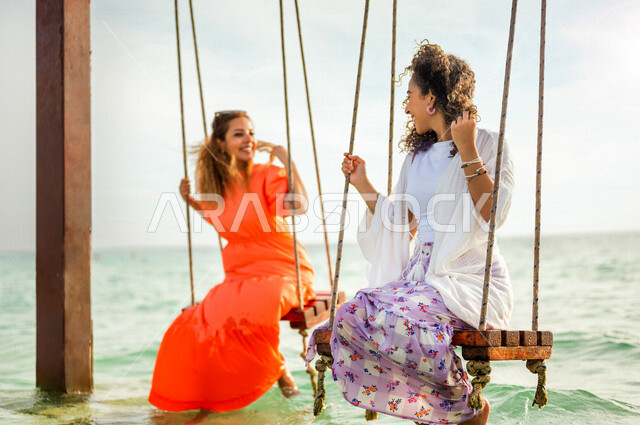 صديقتان عربيتان خليجيتان سعوديتان تقضيان وقت ممتع على الارجوحة المعلقة فوق البحر، بحر جدة السياحي، الاستمتاع بالاجازة الصيفية، ايماءات وجه ويدين تدل على السعادة، سياحة وإستجمام، انشطة ترفيهية، العطلة الصيفية، أزياء صيفية