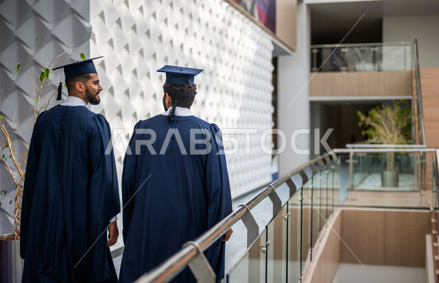 صوره من الخلف لطالبان عربيان سعوديان خليجيان يرتديان العباءة وقبعة التخرج، التجول داخل مبنى الجامعه, الاحتفال بالتخرج من الجامعة ، حفل تخرج ، لباس التخرج ، مبنى الحرم الجامعي, جامعات أكاديميه في المملكه العربيه السعوديه, كليات وجامعات متطوره