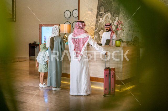 صورة من الخلف لعائلة عربية خليجية سعودية تقف امام مكتب موظفين الإستقبال في فندق سياحي، اتمام إجراءات الحجز والدخول لدى مكتب استقبال العملاء في الفندق، خدمة العملاء، حقيبة سفر حمراء، سياحة وفندقة، رحلة عائلية سعيدة، فنادق فاخرة في السعودية