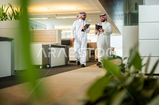 موظفان عربيان سعوديان خليجيان داخل مقر الشركة، يرتديان الزي السعودي التقليدي، مهن و وظائف سعودية، شركة سعودية، رجال أعمال، الحوارات والنقاشات، بيئة العمل، دردشة عمل، متابعة ومناقشة الأعمال، زملاء عمل، مناقشة ومتابعة الأهداف،
