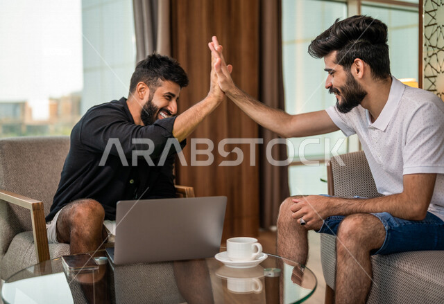 رجلان عربيان خليجيان سعوديان يستخدمان جهاز الحاسوب المحمول ويتناقشان بالعمل، اجتماع زملاء عمل، إتفاق عمل، الوحده والعمل الجماعي، صفقات عمل، كوب قهوة، مشروبات ساخنة، ايماءات وجه سعيدة، سياحة وفندقة، غرفة فندقية