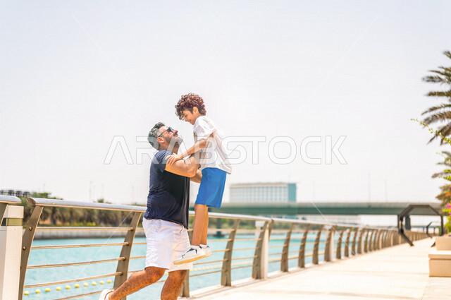 رجل عربي خليجي سعودي يقضي وقت ممتع برفقة إبنه على ممشى كورنيش جدة، كورنيش جدة السياحي، واجهة جدة البحرية، أماكن سياحية ترفيهية بالمملكة العربية السعودية، الأب يحمل إبنه إلى الأعلى، أسرة سعودية