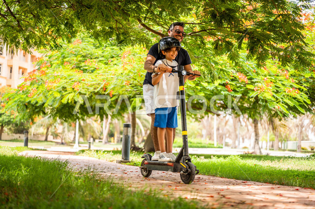 رجل عربي خليجي سعودي يقضي وقت ممتع بقيادة السكوتر الكهربائي مع إبنه في إحدى الحدائق السياحية الترفيهية في المملكة العربية السعودية، مشاركة الأبناء الأنشطة الترفيهية، تعابير تدل على السعادة، قضاء أوقات عائلية ممتعة، حدائق عامة، خلفية الطبيعة