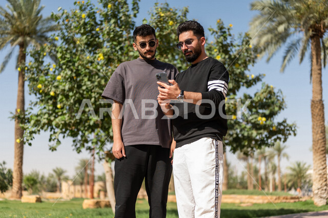 صورة مقربة لشابان عربيان خليجيان سعوديان يرتديان النظارات الشمسية ويستخدمان الهاتف المحمول، نزهه في الهواء الطلق، استخدام البرامج والتطبيقات الالكترونية، التسوق الالكتروني، التواصل الاجتماعي، تصفح المواقع الالكترونية، حدائق ومنتزهات