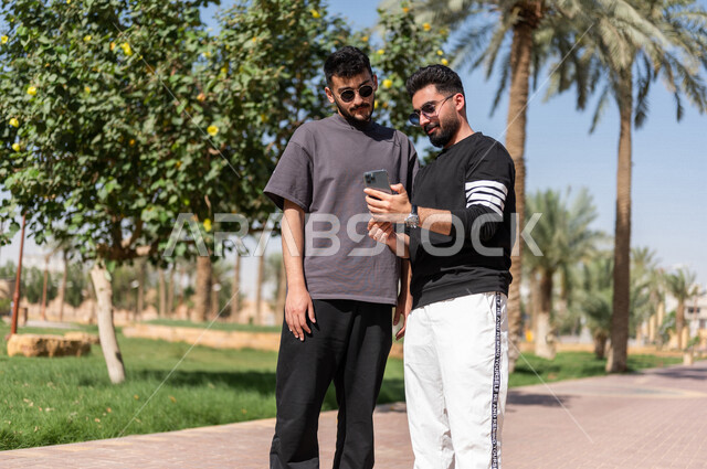 صورة مقربة لشابان عربيان خليجيان سعوديان يرتديان النظارات الشمسية ويستخدمان الهاتف المحمول، نزهه في الهواء الطلق، استخدام البرامج والتطبيقات الالكترونية، التسوق الالكتروني، التواصل الاجتماعي، تصفح المواقع الالكترونية، حدائق ومنتزهات