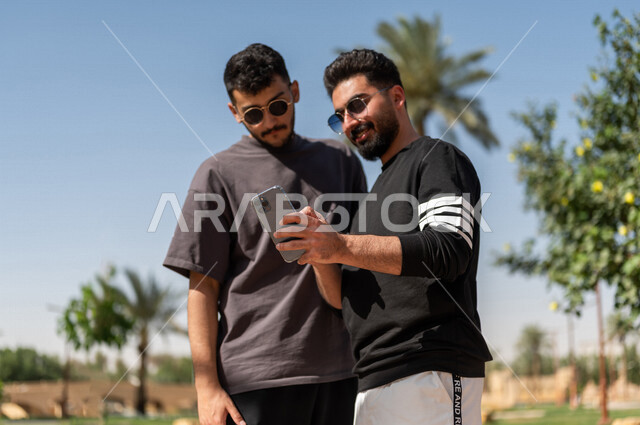 صورة مقربة لشابان عربيان خليجيان سعوديان يرتديان النظارات الشمسية ويستخدمان الهاتف المحمول، التنزه في الهواء الطلق، استخدام البرامج والتطبيقات الالكترونية، التسوق الالكتروني، التواصل الاجتماعي، تصفح المواقع الالكترونية، ادمان الانترنت، حدائق ومنتزهات
