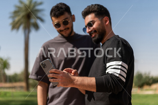 صورة مقربة لشابان عربيان خليجيان سعوديان يرتديان النظارات الشمسية ويستخدمان الهاتف المحمول، التنزه في الهواء الطلق، استخدام البرامج والتطبيقات الالكترونية، التسوق الالكتروني، التواصل الاجتماعي، تصفح المواقع الالكترونية، ادمان الانترنت، حدائق ومنتزهات