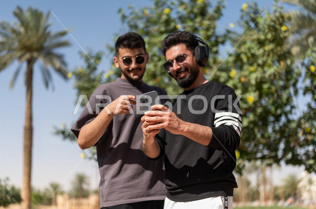 صورة مقربة لشابان عربيان خليجيان سعوديان يرتديان النظارات الشمسية ويستخدمان الهاتف المحمول، استخدام البرامج والتطبيقات الالكترونية، شاب يرتدي سماعات الرأس الصوتية، الإستماع إلى الموسيقى، إستخدام تقنية الهيدفون، الإصغاء والإستمتاع، حدائق ومنتزهات