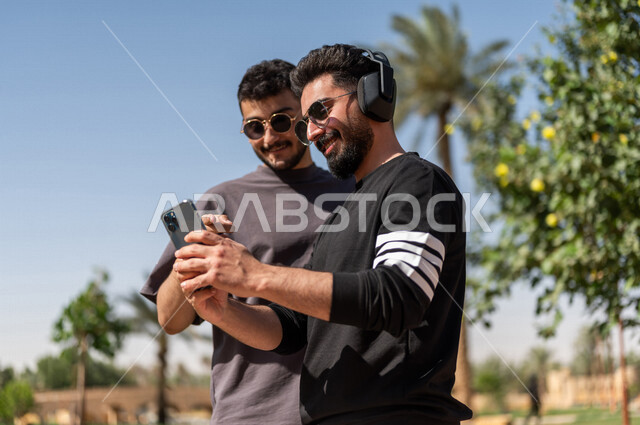 صورة مقربة لشابان عربيان خليجيان سعوديان يرتديان النظارات الشمسية ويستخدمان الهاتف المحمول، استخدام البرامج والتطبيقات الالكترونية، شاب يرتدي سماعات الرأس الصوتية، الإستماع إلى الموسيقى، إستخدام تقنية الهيدفون، الإصغاء والإستمتاع، حدائق ومنتزهات