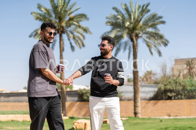 شابان عربيان خليجيان سعوديان يتقابلان في احد الحدائق العامة ويتصافحان بابتسامة، ممارسة رياضة المشي الهواء الطلق مع احتساء كوب من القهوة، التصافح بحرارة، تعارف وصداقات، السلام والترحيب، طريق ممشى في الحدائق والمنتزهات، نزهه في الهواء الطلق