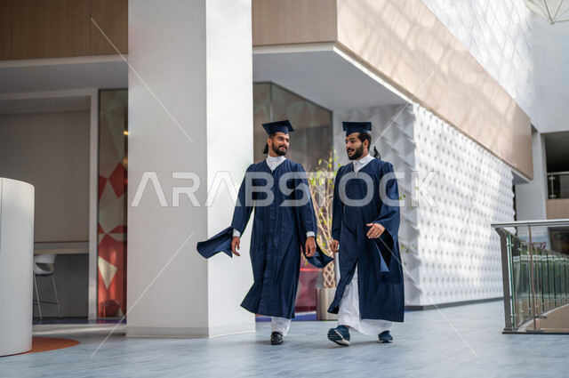 طالبان عربيان سعوديان خليجيان يرتديان العباءة وقبعة التخرج، التجول داخل مبنى الجامعه, الاحتفال بالتخرج من الجامعة ، حفل تخرج ، لباس التخرج ، مبنى الحرم الجامعي, ايماءات تدل على السعاده بالتفوق والنجاح, جامعات أكاديميه في السعوديه, كليات وجامعات متطوره