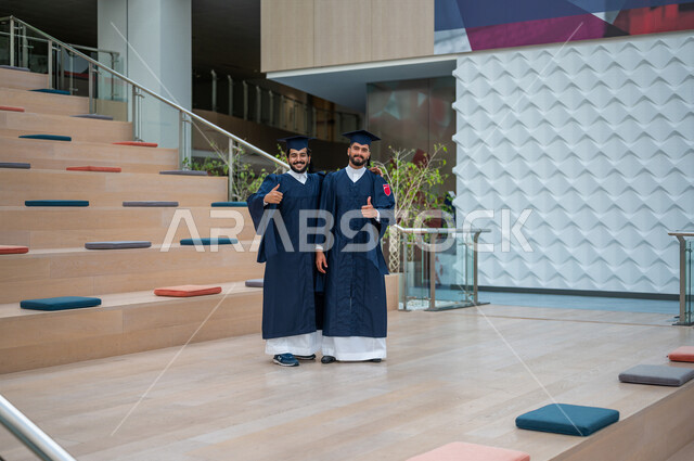 طالبان عربيان سعوديان خليجيان يرتديان العباءة وقبعة التخرج ، الاحتفال بالتخرج من الجامعة ، حفل تخرج ، لباس التخرج ، مبنى الحرم الجامعي, ايماءات تدل على السعاده بالتفوق والنجاح, جامعات أكاديميه في المملكه العربيه السعوديه, كليات وجامعات متطوره