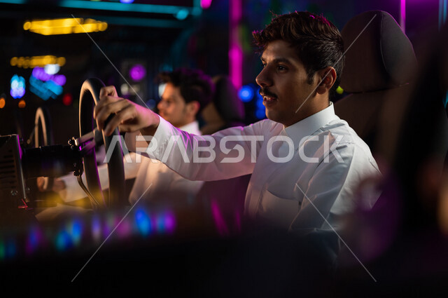 شابان مراهقان عربيان خليجيان سعوديان في متجر الألعاب الإلكترونية، لعبة سيارات سباق، الجلوس خلف عجلة مقود السيارة (يد التحكم)، تعابير تدل على الإندماج، ألعاب الفيديو الإلكترونية، تكنولوجيا الألعاب، أنشطة ترفيهية، قضاء وقت الفراغ بألعاب الفيديو مع الأصدقاء