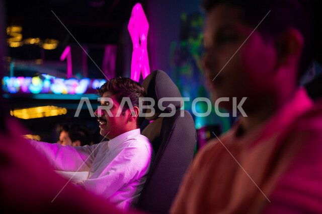 شابان مراهقان عربيان خليجيان سعوديان في متجر الألعاب الإلكترونية، لعبة سيارات سباق، الجلوس خلف عجلة مقود السيارة (يد التحكم)، تعابير تدل على الإندماج، ألعاب الفيديو الإلكترونية، تكنولوجيا الألعاب، أنشطة ترفيهية، قضاء وقت الفراغ بألعاب الفيديو مع الأصدقاء