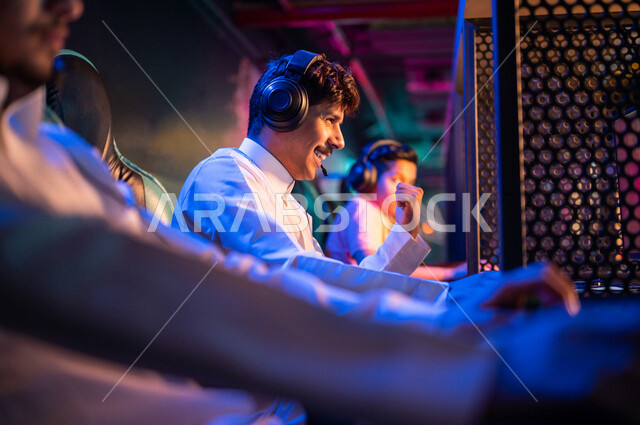 شباب عرب مراهقين خليجيين سعوديين في متجر الألعاب الإلكترونية، يرتديان سماعة الرأس ويلعبان ألعاب الفيديو الإلكترونية، إيماءات تدل على الإندماج والتفاعل، أنشطة ترفيهية، ألعاب الكمبيوتر الترفيهية، قضاء وقت الفراغ بألعاب الفيديو مع الأصدقاء، اللعب الجماعي