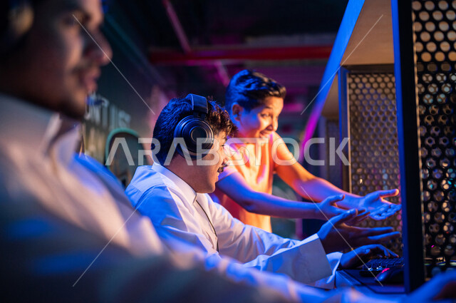شباب عرب مراهقين خليجيين سعوديين في متجر الألعاب الإلكترونية، يرتديان سماعة الرأس ويلعبان ألعاب الفيديو الإلكترونية، إيماءات تدل على الإندماج والتفاعل، أنشطة ترفيهية، ألعاب الكمبيوتر الترفيهية، قضاء وقت الفراغ بألعاب الفيديو مع الأصدقاء، اللعب الجماعي