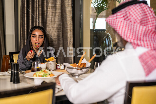زوجان عربيان سعوديان خليجيان يجلسان في مطعم، مائدة طعام مليئة بأصناف متنوعة من المأكولات والمشروبات الشهية، نقاشات وحوارات عائلية، تعابير تدل على السعادة، قضاء أوقات عائلية ممتعة، سفرة طعام، اجواء عائلية سعيدة، مطاعم وكافيهات راقية في السعودية
