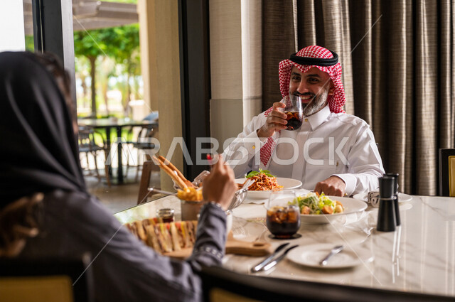 زوجان عربيان سعوديان خليجيان يجلسان في مطعم، مائدة طعام مليئة بأصناف متنوعة من المأكولات والمشروبات الشهية، نقاشات وحوارات عائلية، تعابير تدل على السعادة، قضاء أوقات عائلية ممتعة، سفرة طعام، اجواء عائلية سعيدة، مطاعم وكافيهات راقية في السعودية