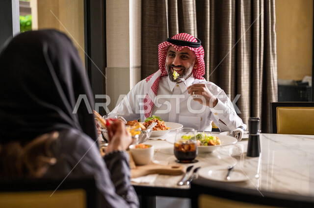 زوجان عربيان سعوديان خليجيان يجلسان في مطعم، مائدة طعام مليئة بأصناف متنوعة من المأكولات والمشروبات الشهية، نقاشات وحوارات عائلية، تعابير تدل على السعادة، قضاء أوقات عائلية ممتعة، سفرة طعام، اجواء عائلية سعيدة، مطاعم وكافيهات راقية في السعودية